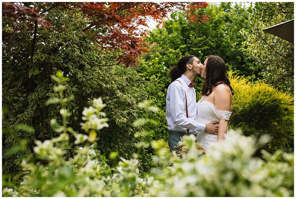 A couple embraces in a lush garden, sharing a kiss surrounded by vibrant greenery and flowers, capturing a moment of love and intimacy.