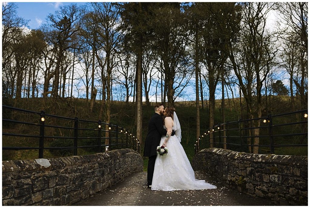 A bride and groom share a kiss on a picturesque bridge surrounded by trees, with a serene sky in the background.