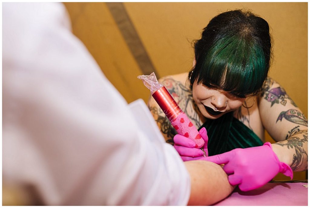 A tattoo artist with green hair and pink gloves focuses on tattooing a client's arm while using a colorful tattoo gun.