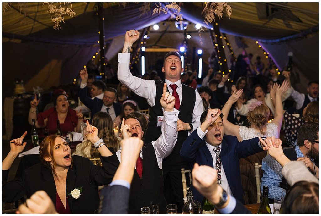 Guests joyfully singing and cheering with raised hands in a lively indoor setting filled with warm lighting and decorations.