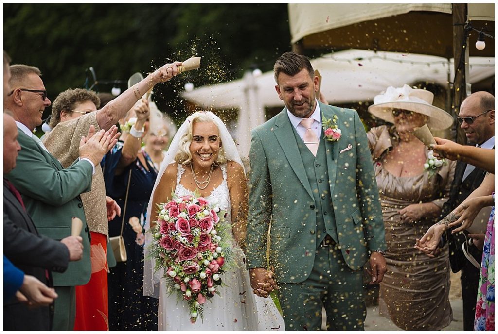 Wedding Celebration Filled with Joy:::

A happy bride and groom walk hand-in-hand, surrounded by guests tossing confetti in a vibrant outdoor setting. The bride holds a beautiful bouquet of pink roses, and both are smiling broadly as they celebrate their special day.