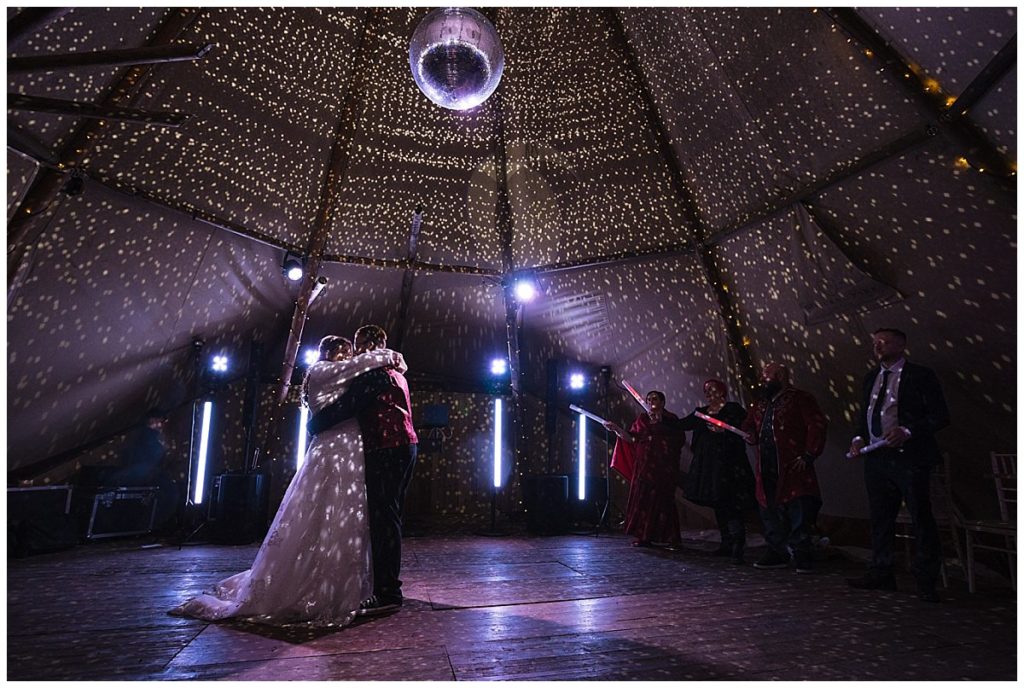 A couple embraces during their first dance in a tent adorned with shimmering lights and a disco ball, surrounded by joyful guests.
