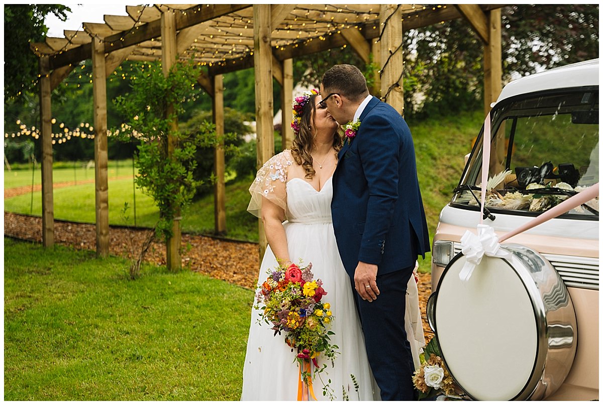 A couple shares a kiss under a wooden pergola adorned with fairy lights, surrounded by lush greenery and colorful flowers, on their special day.