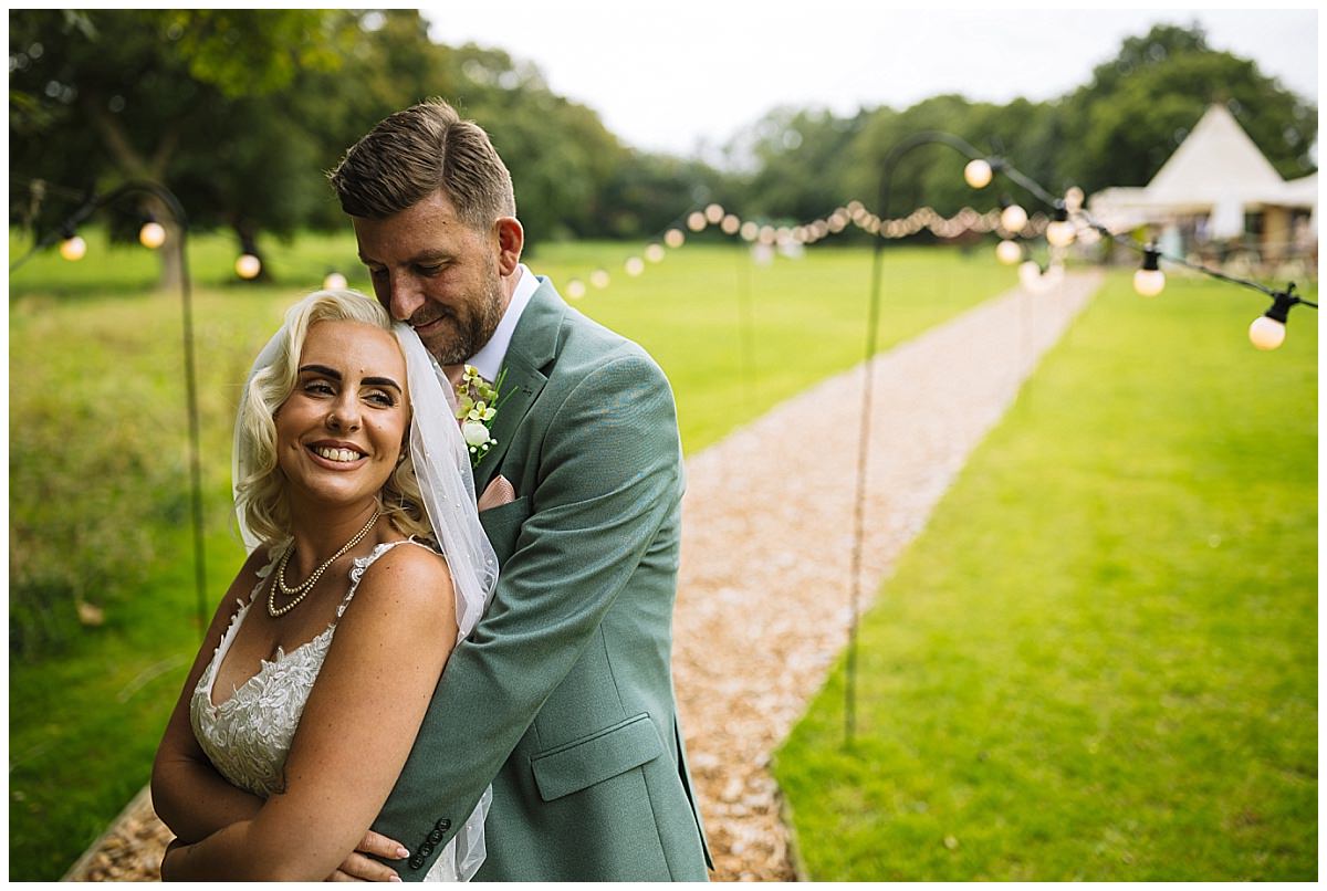 Happy couple embracing in a picturesque outdoor setting, adorned with string lights and a scenic path at a tipi wedding
