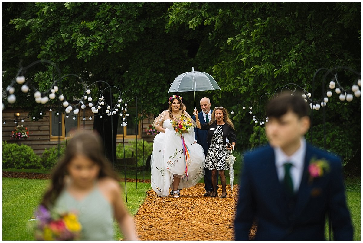 A bride adorned with flowers walks down an aisle accompanied by guests, while a child in the foreground strolls past, set against a backdrop of greenery and decorative lights.