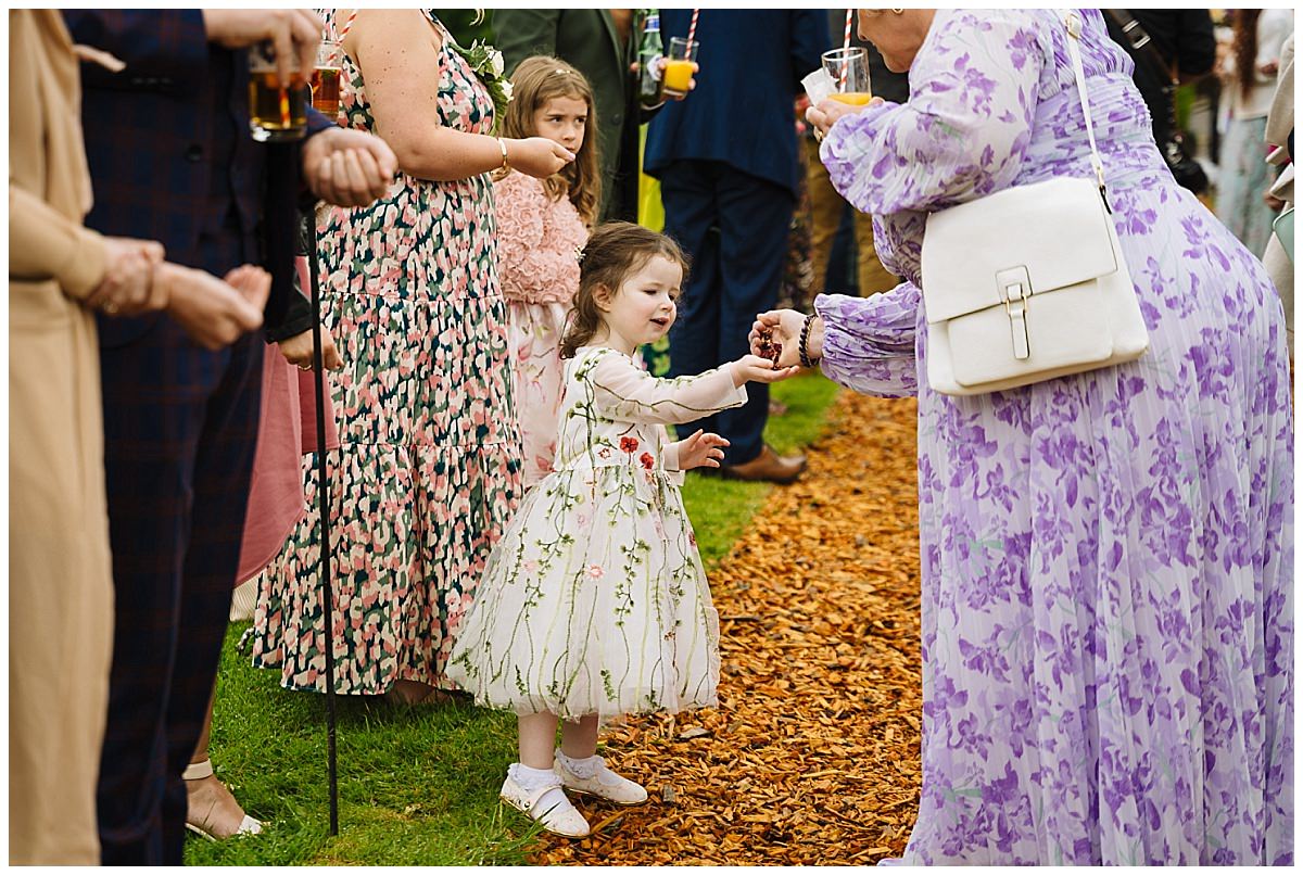A young girl in a floral dress reaches out to receive a treat from a woman in a lavender dress while surrounded by guests at a festive event.