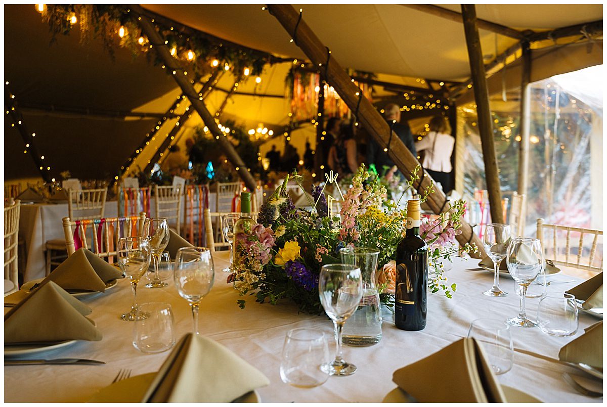 A beautifully arranged table in a tipi, featuring floral centerpieces, wine bottles, and glassware, set for a celebratory occasion with warm lighting in the background.