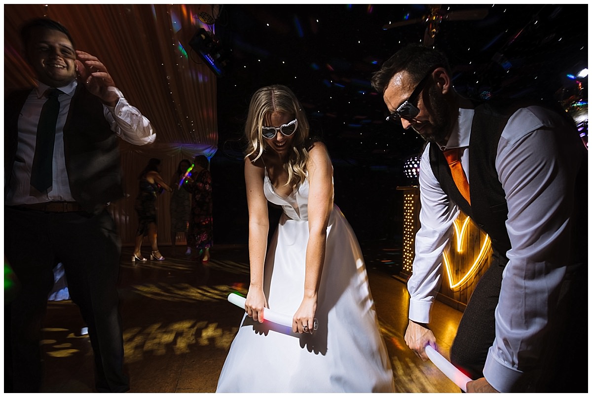 A bride and two guests enjoying a lively dance floor, holding glow sticks and wearing sunglasses, in a festive atmosphere filled with colorful lights.