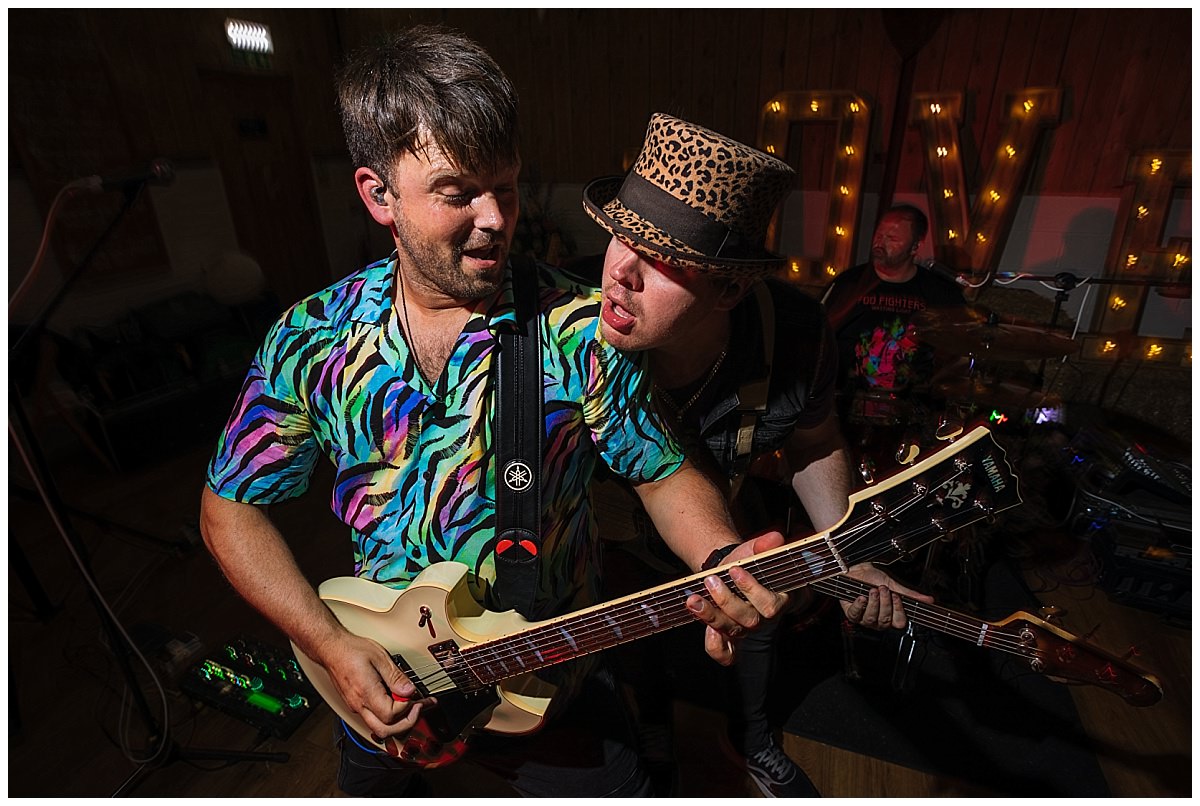 Two male musicians performing passionately on stage, one playing an electric guitar and the other singing, both dressed in vibrant, colorful outfits.