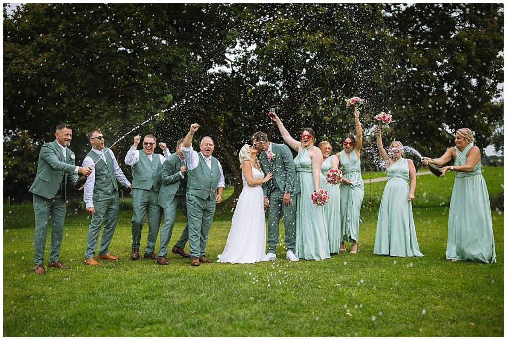 A joyful wedding party celebrating with champagne, as the bride and groom share a kiss amidst cheering friends in matching green attire.