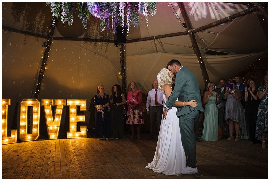 A bride and groom share a romantic first dance in a beautifully decorated venue, with a large illuminated "LOVE" sign in the background and guests celebrating around them.
