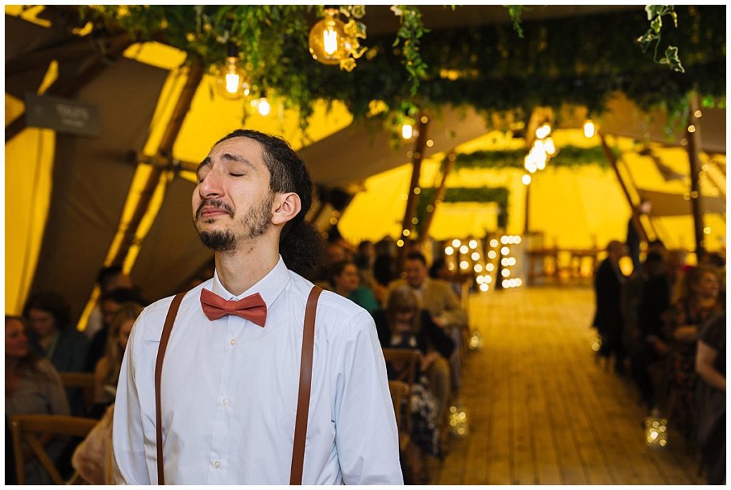 A man in a white shirt and bow tie stands with closed eyes, expressing emotion in a decorated tent filled with guests, illuminated by warm lights.