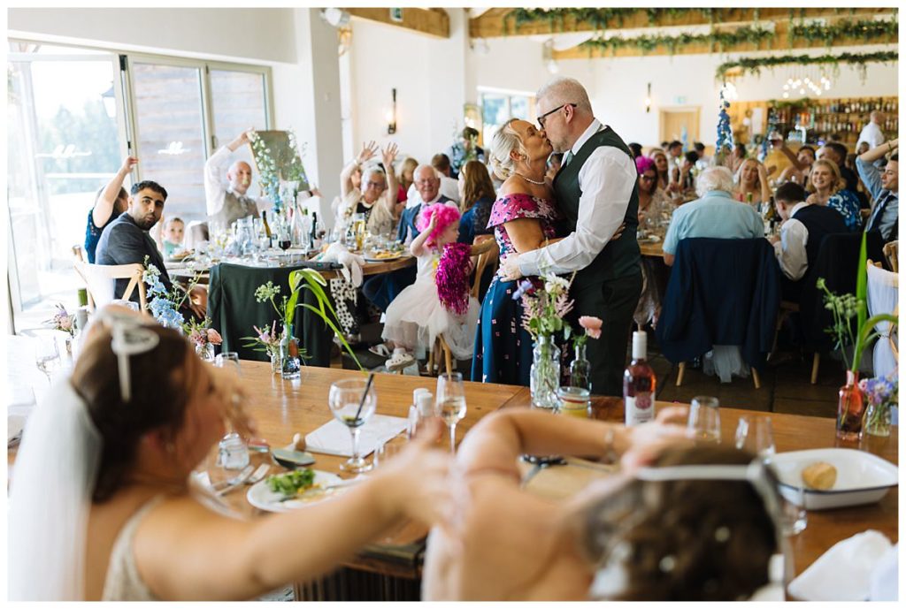 A joyful couple shares a kiss during their wedding reception, surrounded by cheering guests and elegantly set tables.