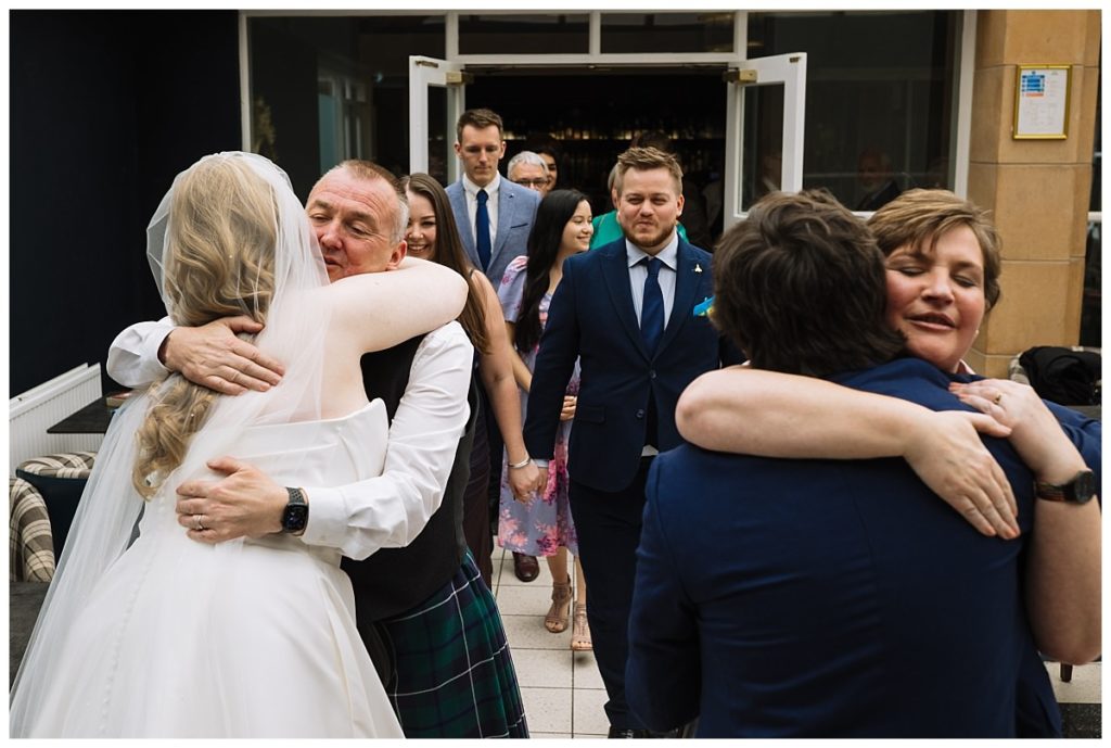 A joyful wedding scene capturing guests hugging, with the bride in a white dress and veil, surrounded by family and friends in formal attire.