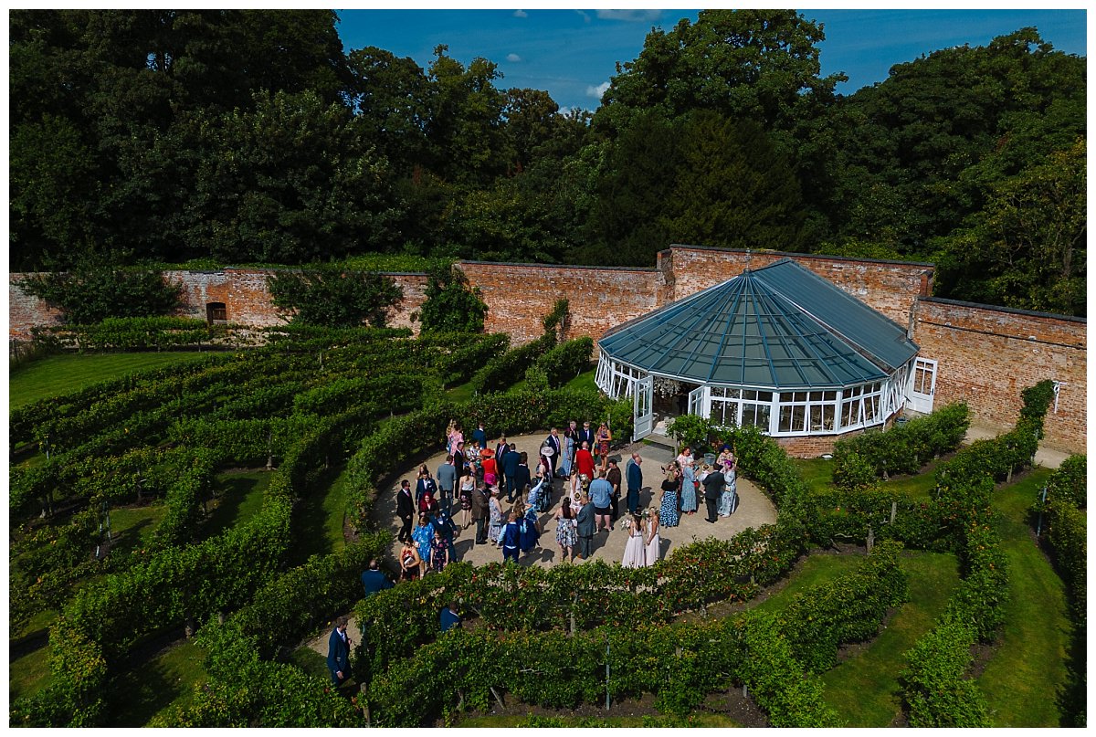 Aerial view of a group of people socializing in a circular garden surrounded by lush green vines and a glassed conservatory in the background.