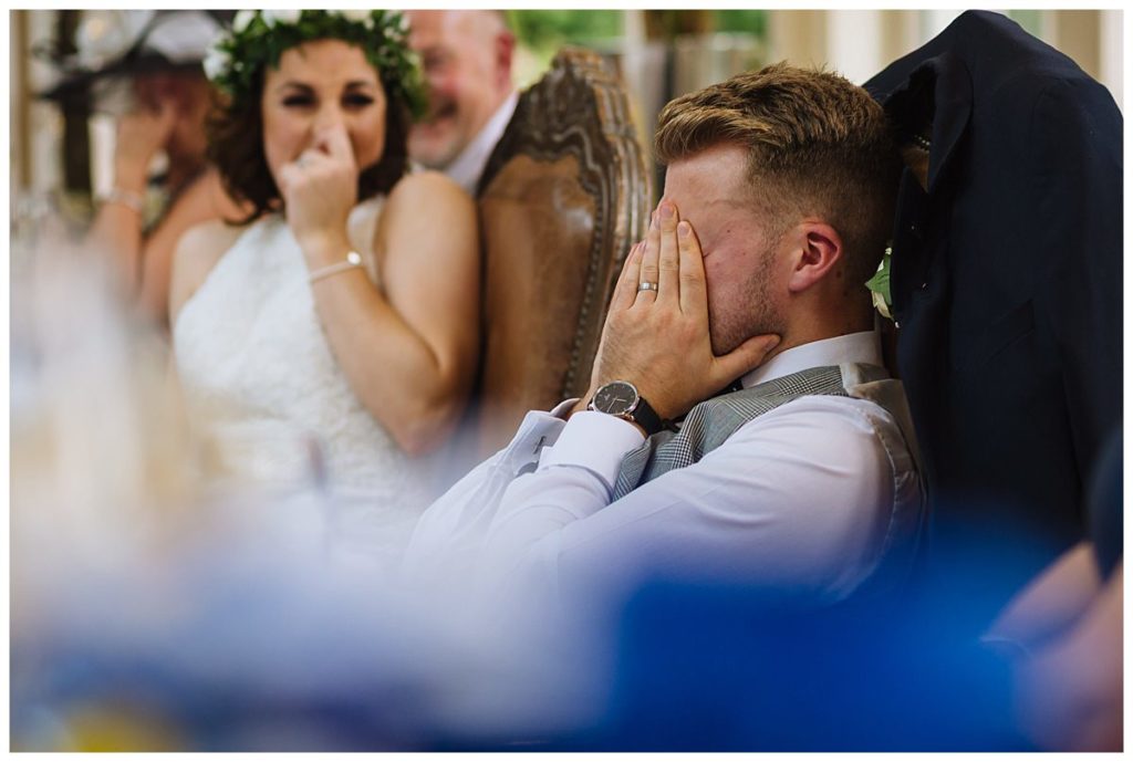 A man sits with his hands over his face, eliciting a moment of laughter and surprise, while a woman in a white dress and floral crown reacts joyfully in the background.