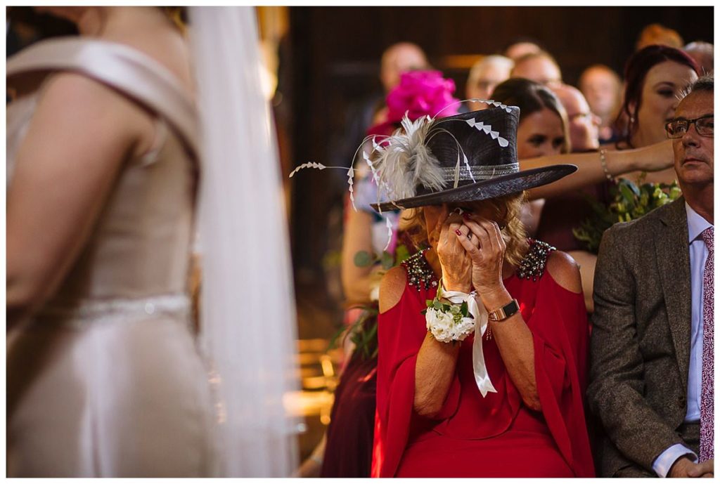 A woman in a red dress and large hat wipes away tears during a wedding ceremony, capturing a heartfelt reaction amidst a seated audience.