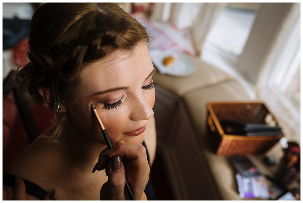 A close-up of a woman having her eye makeup applied, with a makeup brush in hand and beauty products visible in the background.