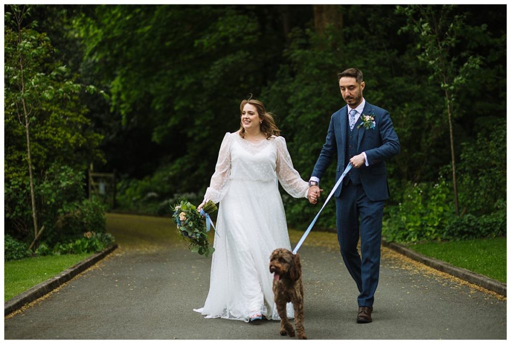 A newly married couple strolls hand-in-hand along a pathway with their dog, surrounded by lush greenery.