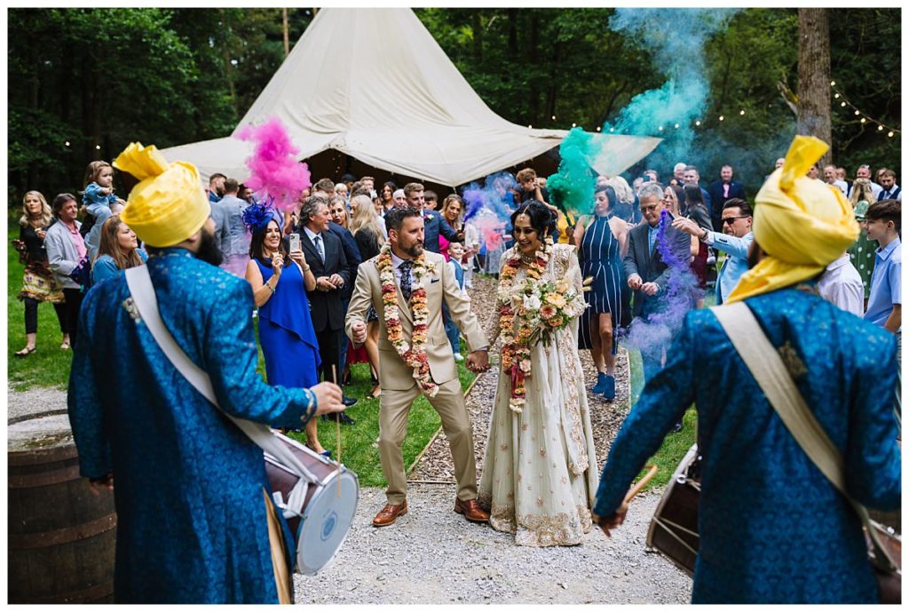 A joyous couple leads a wedding procession amid colorful smoke, surrounded by smiling guests at an outdoor venue.