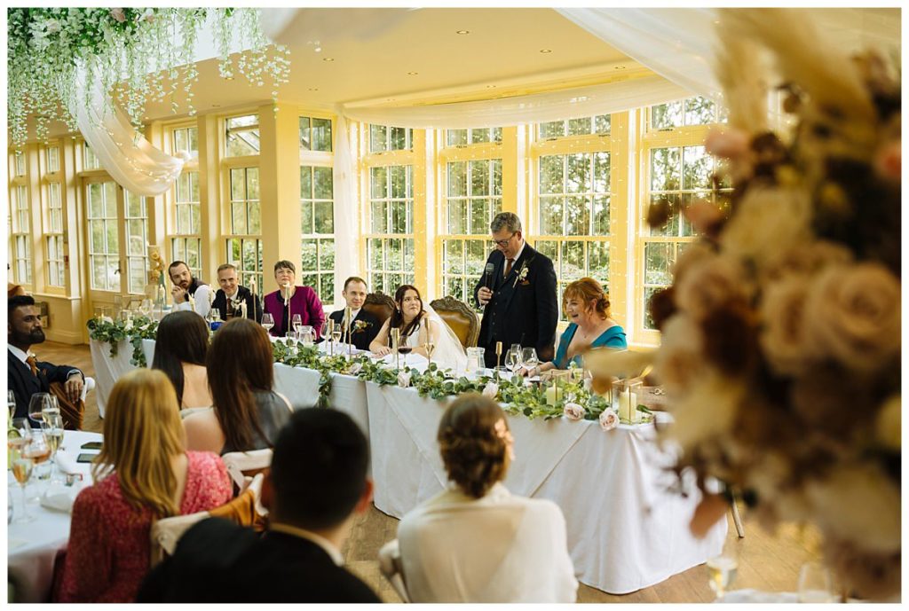 A joyous wedding reception scene featuring guests enjoying speeches at a beautifully decorated table with greenery and floral arrangements, bathed in warm natural light.