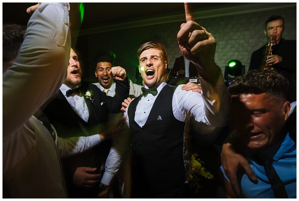 A lively group of men celebrating at a wedding reception, dancing and singing with vibrant lighting in the background.