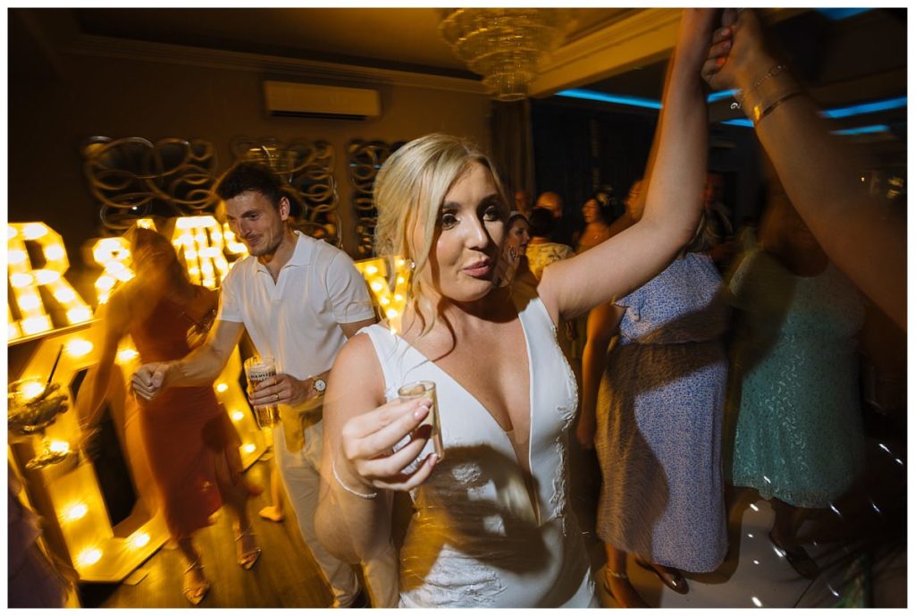 A woman in a white dress raises a shot glass while dancing at a lively wedding reception, surrounded by guests in festive attire and illuminated by glowing decorations.