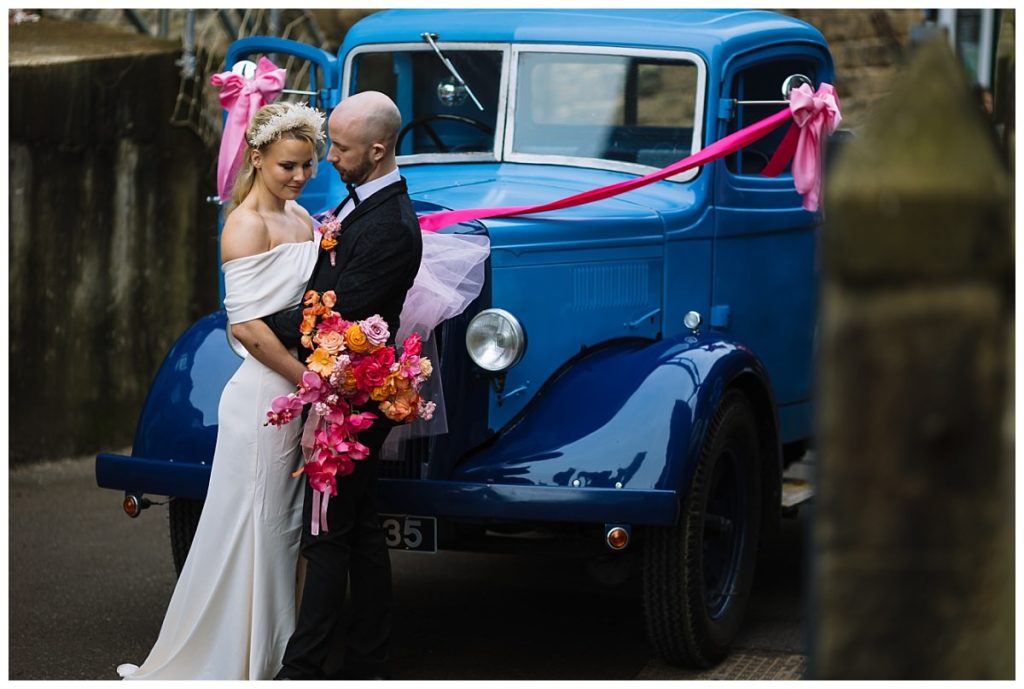A couple in formal attire poses lovingly in front of a classic blue vintage car, adorned with pink ribbons and a vibrant bouquet.