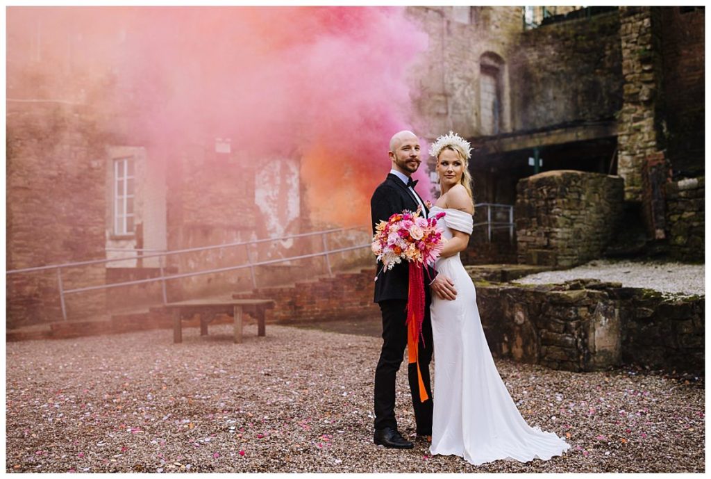 A couple poses together in a romantic setting, surrounded by pink smoke, showcasing the bride's elegant dress and vibrant bouquet.