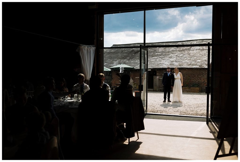 A bride and groom standing at an open doorway, framed by shadows and light, with guests seated at tables in the foreground.