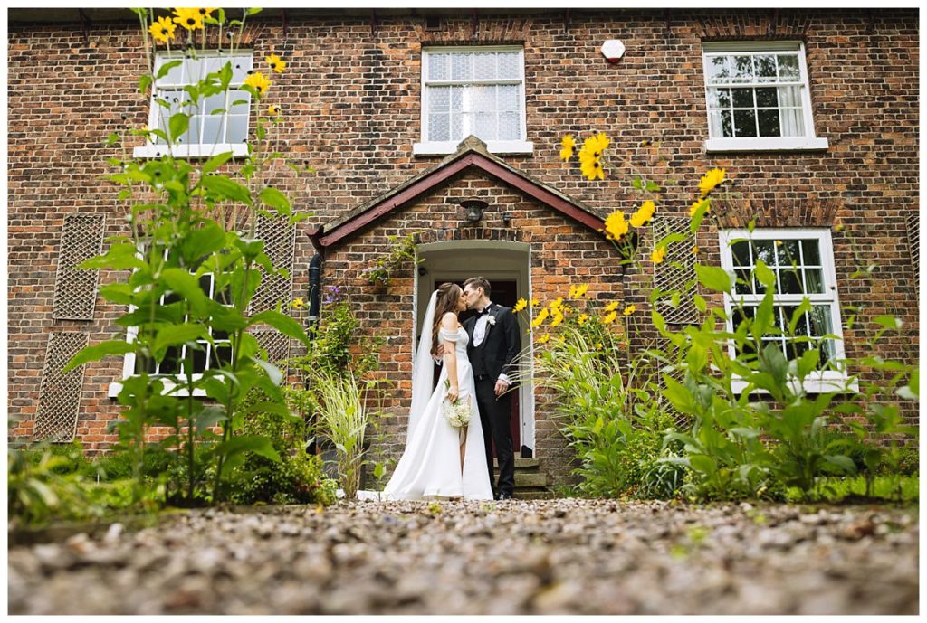 A couple shares a kiss, surrounded by lush greenery and vibrant flowers at the entrance of a charming brick house.