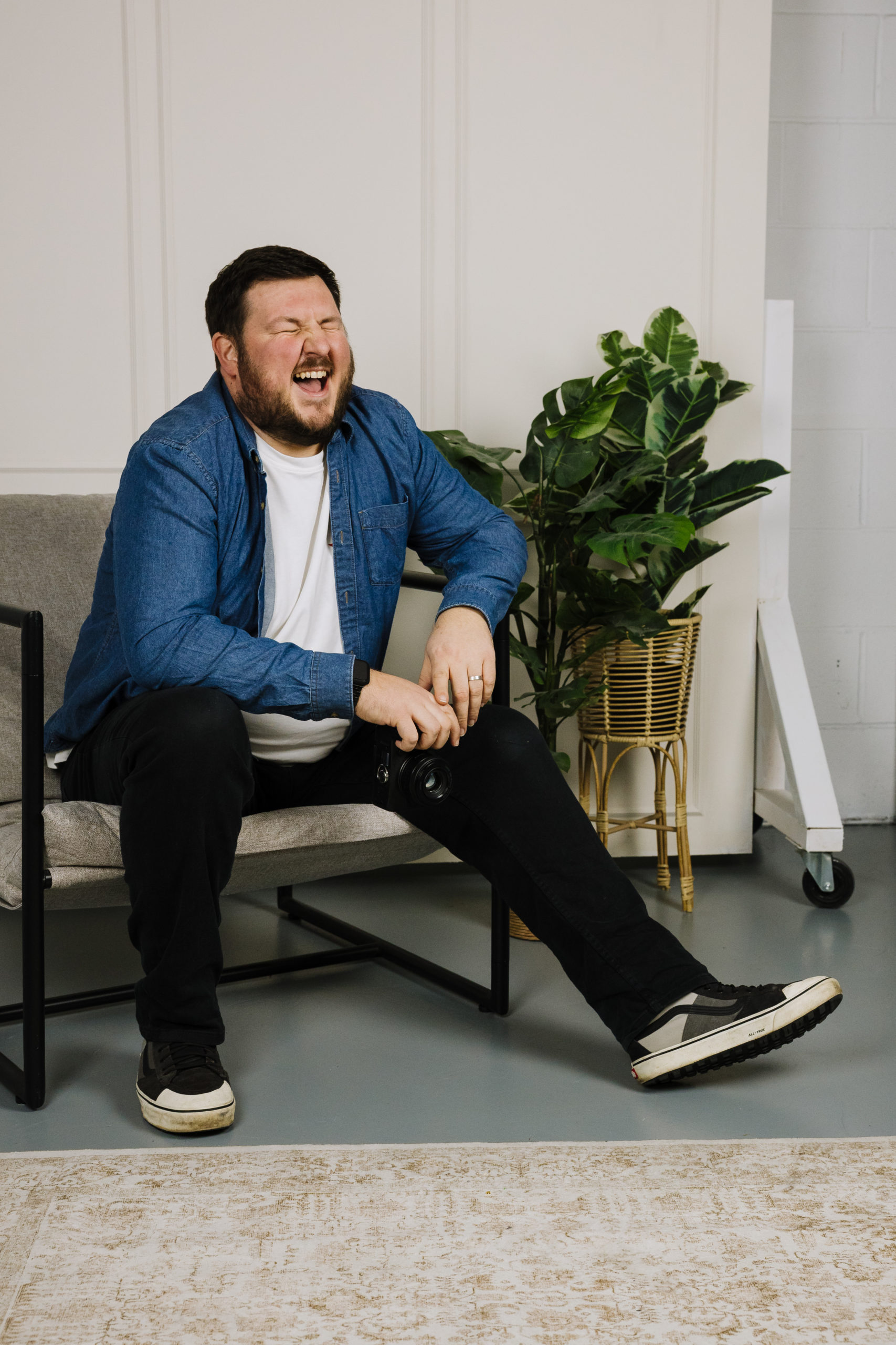 Nik Bryant Photography sitting on a chair, laughing joyfully while holding a camera, surrounded by indoor plants and a stylish interior.